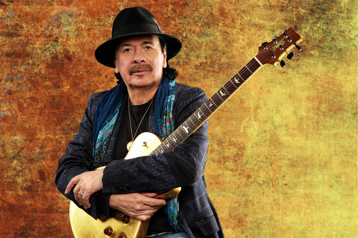 Carlos Santana holding a gold electric guitar against a warm gold background